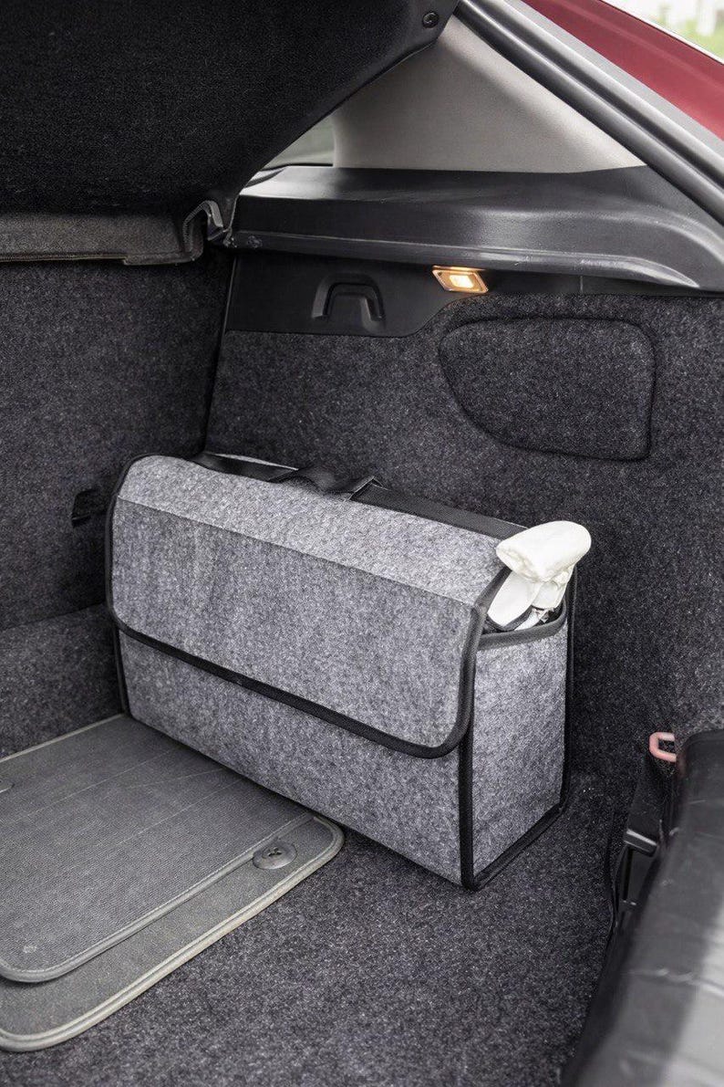 May include: A gray felt car trunk organizer with a black trim and handle, placed inside a car trunk. The organizer is rectangular with a flap closure and contains white items. A gray car mat is visible.