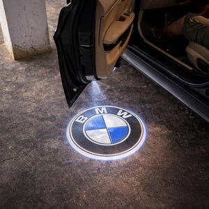 May include: A black car door is open, illuminating the ground with a BMW logo projection. The logo is a white and blue emblem within a black circle, with the letters "BMW" at the top. The car's interior is visible.