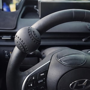 Puede incluir: Un pomo de volante negro con temática de béisbol está sujeto al volante de un coche. El pomo es redondo, con detalles cosidos, que se asemejan a una pelota de béisbol. El volante es negro, con controles integrados.