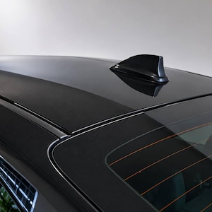 May include: A glossy black car roof featuring a sleek, aerodynamic shark fin antenna positioned toward the rear. The roof panel transitions into the rear window, which displays thin, horizontal orange defroster lines across the dark glass surface.