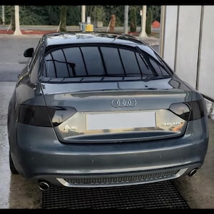 Op de afbeelding: Grijze Audi A5 coupé, gezien van achteren, geparkeerd op een nat oppervlak. De auto heeft getinte ramen en zwart gemaakte achterlichten. Het Audi-logo is zichtbaar, samen met de tekst "A5" en "3.0 TDI".