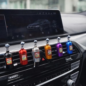 Può includere: Una fila di profumatori per auto colorati a forma di bottiglie NOS. Le bottiglie sono nere, rosse, argentate, arancioni, viola e blu, ognuna con un tappo argentato e un'etichetta "NOS". Sono posizionati sul cruscotto di un'auto.