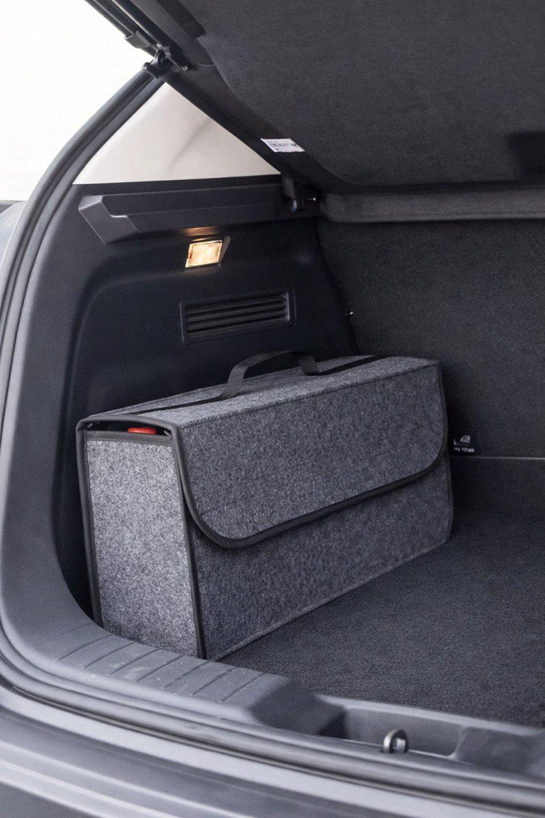May include: A gray felt car trunk organizer with a black handle and trim, inside the trunk of a car. The organizer has a flap closure and a rectangular shape. The car's interior is dark gray.