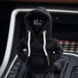 以下が含まれることがあります： 白い紐とフードが付いた、黒いパーカーの車のギアシフトカバー。車のギアシフトレバーにかぶせるように設計されています。ギアシフトレバーには「N」、「D」、「M」、「+」の文字が表示されています。