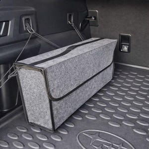 May include: A gray felt storage container with black trim and a black strap, secured in a car trunk. The container is rectangular and sits on a textured, dark gray mat. The trunk interior is dark gray.