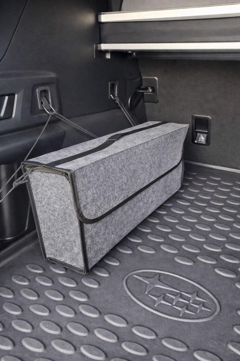 May include: A gray felt storage container with black trim and a black strap, secured in a car trunk. The container is rectangular and sits on a textured, dark gray mat. The trunk interior is dark gray.