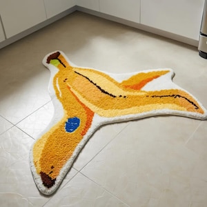 May include: A novelty banana-shaped rug with a white border. The rug is in shades of yellow, orange, and brown, with a blue spot. The rug is made of a soft, textured material and is designed to resemble a peeled banana. It would look great in a kitchen.