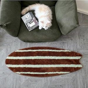 May include: An oval-shaped rug with a fish-tail end, featuring horizontal stripes in alternating shades of dark brown and light mint green. The rug has a soft, textured pile and is positioned on a light grey floor with a herringbone pattern.