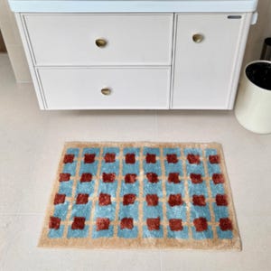 Può includere: Un tappetino da bagno rettangolare con un motivo a griglia di rettangoli azzurri e piccoli quadrati testurizzati in rilievo color ruggine. Il tappetino ha un bordo color marrone chiaro ed è posizionato su un pavimento chiaro davanti a un mobile bianco.