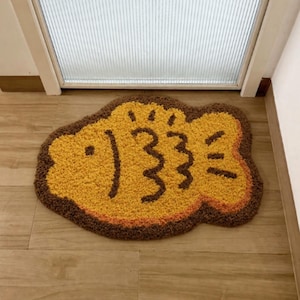 May include: A tufted rug shaped like a fish, featuring a bright yellow center with dark brown outlines and detailing. The rug has a textured, shaggy pile surface and a distinct fish silhouette with a rounded body, fins, and a tail section.