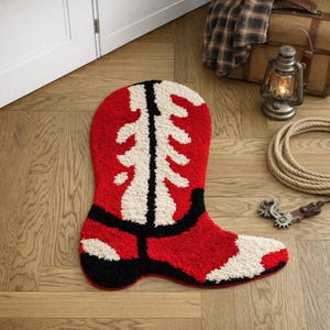 May include: A red, white, and black cowboy boot-shaped rug. The rug has a textured, tufted appearance. The boot design features a white shaft with a black outline and a red foot. A western-themed decorative item.