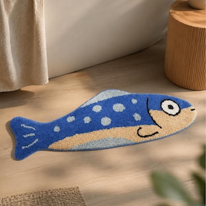 May include: A fish-shaped bath mat featuring a textured blue body with light blue polka dots, a beige face with a large white eye and black pupil, and a beige underbelly. The rug has a soft, tufted pile surface and a distinct fish silhouette.