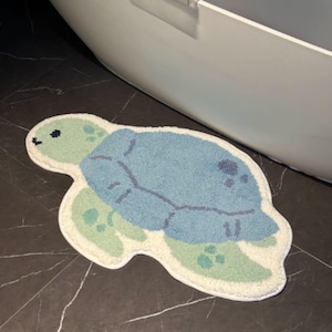 Op de afbeelding: Een getufte badmat in de vorm van een zeeschildpad met een lichtgroene kop en flippers, een blauw schild met donkerblauwe stippen en een witte rand. Het kleed heeft een getextureerd, pluche oppervlak en is ontworpen in de vorm van een schildpad.