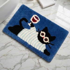 Op de afbeelding: Een rechthoekige blauwe badmat met een zwarte kat die een lichtblauw en wit gestreept shirt en een ronde bril draagt. De kat houdt een glas rode wijn vast met zijn poot. Het kleed heeft een getextureerde, getufte oppervlakteafwerking.