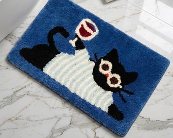 May include: A rectangular blue bath mat featuring a black cat wearing a light blue and white striped shirt and round glasses. The cat is depicted holding a glass of red wine with its paw. The rug has a textured, tufted surface finish.