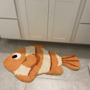 Peut inclure: Un tapis en forme de poisson avec un poil touffeté texturé dans des tons orange, crème et blanc. Le motif comprend un détail d'œil noir et blanc proéminent, des rayures distinctes sur le corps et une nageoire caudale en éventail avec des lignes orange.