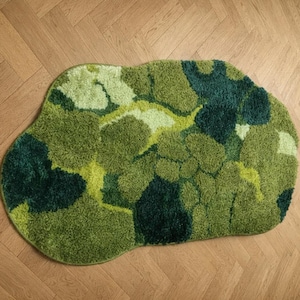 Könnte beinhalten: Ein unregelmäßig geformter Teppich mit einer hochflorigen, getufteten Textur. Der Teppich zeigt ein abwechslungsreiches Muster aus verschiedenen Grüntönen, das von tiefem Waldgrün und dunklem Smaragd bis zu hellem Limettengrün und Salbei reicht.