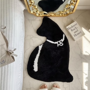 May include: A black cat-shaped rug with a white collar and a white stripe down the side. The rug is on a tiled floor, with a book titled "Madness, Rack, and Honey" nearby. A gold-framed mirror reflects the rug.
