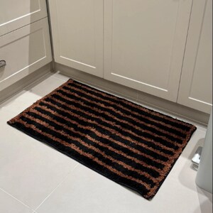 May include: A rectangular floor mat featuring a repeating pattern of horizontal stripes in alternating black and copper-brown colours. The mat has a textured, shaggy pile surface and is positioned on a light-coloured tiled floor in front of cabinets.