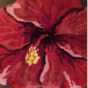May include: A large, plush rug in the shape of a hibiscus flower. The rug features shades of red, pink, and burgundy, with a yellow and white center. The floral design is detailed and textured, creating a soft and inviting aesthetic.