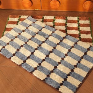 Pode incluir: Dois tapetes retangulares de pelo alto com um padrão de grade repetitivo. O tapete superior apresenta segmentos retangulares em azul claro e creme separados por detalhes em castanho escuro. O tapete inferior exibe um padrão semelhante em laranja ferrugem e creme.