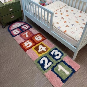 May include: A rectangular hopscotch rug featuring a multi-colored design with white numbers one through ten printed in individual squares. The rug has a textured pile and is positioned on the floor next to a light blue wooden toddler bed.