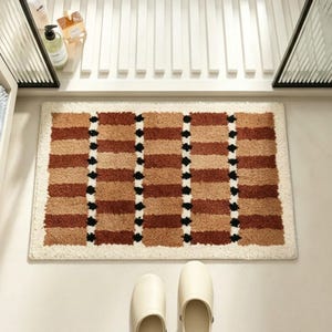 May include: A rectangular bath mat featuring a textured, shaggy pile in a repeating pattern of alternating brown and tan horizontal stripes, separated by vertical rows of small black dots, all bordered by a solid cream-coloured frame.