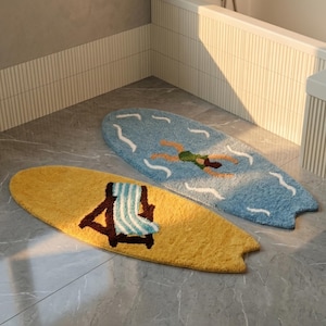Peut inclure: Deux tapis de bain en forme de planche de surf. L'un est jaune avec une chaise de plage marron et une serviette à rayures bleues et blanches. L'autre est bleu avec un nageur et des détails de vagues blanches. Les deux tapis sont sur un sol carrelé gris.