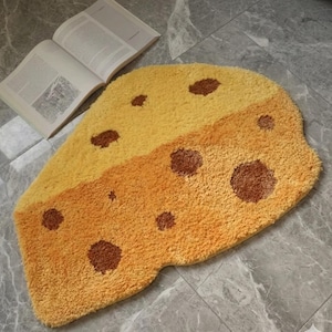 May include: A yellow and orange cheese-shaped rug with brown spots. The rug is made of a soft, plush material. An open book is in the background. This rug is a fun and unique addition to any room.