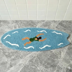 Może przedstawiać: Dywanik w kształcie deski surfingowej w kolorze jasnoniebieskim z tuftowanym wzorem osoby pływającej w ciemnozielonym stroju kąpielowym. Dywanik zawiera białe faliste linie przedstawiające zmarszczki wody i ma klasyczny ogon rybi.
