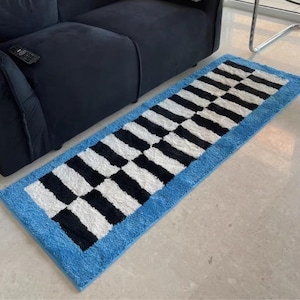 May include: A rectangular runner rug featuring a central pattern of alternating black and white stripes arranged in a grid-like formation, bordered by a solid bright blue frame. The rug has a textured, tufted surface and is placed on a floor.