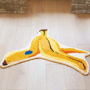Może przedstawiać: Kapryśny dywan w kształcie skórki od banana, z odcieniami żółtego, pomarańczowego i brązowego. Dywan ma białą obwódkę i niebieski akcent. Jest umieszczony na jasnej drewnianej podłodze.