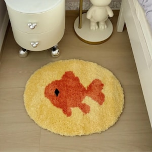 May include: A round, yellow, shaggy-textured rug featuring a centred, orange, fish-shaped design with a small black eye detail. The rug has a thick, plush pile and is positioned on a light-coloured wooden floor surface in a room setting.