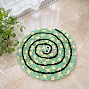 Puede incluir: Una alfombra circular de tejido tufting con un diseño de serpiente enroscada. La serpiente es de color verde claro con manchas amarillas y un contorno negro, con dos ojos blancos y una pequeña lengua rosa visibles en el centro del patrón en espiral.