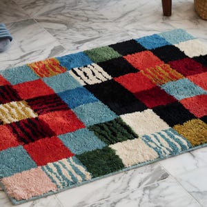May include: A rectangular bath mat featuring a multi-colored checkerboard pattern. The rug is composed of plush, textured squares in shades of red, blue, black, green, yellow, and cream, with some squares displaying a subtle striped texture.