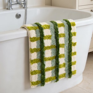 May include: A rectangular bath mat featuring a textured, shaggy pile in a plaid pattern. The design consists of alternating vertical and horizontal stripes in shades of cream, sage green, and vibrant lime green, creating a grid-like appearance.