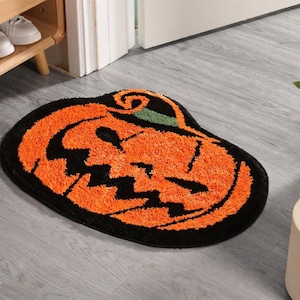Może przedstawiać: Dywan w stylu Halloween w kształcie dyni. Dywan ma pomarańczową twarz dyni z czarnym konturem i zieloną łodygą na czarnym tle. Leży na szarej drewnianej podłodze.