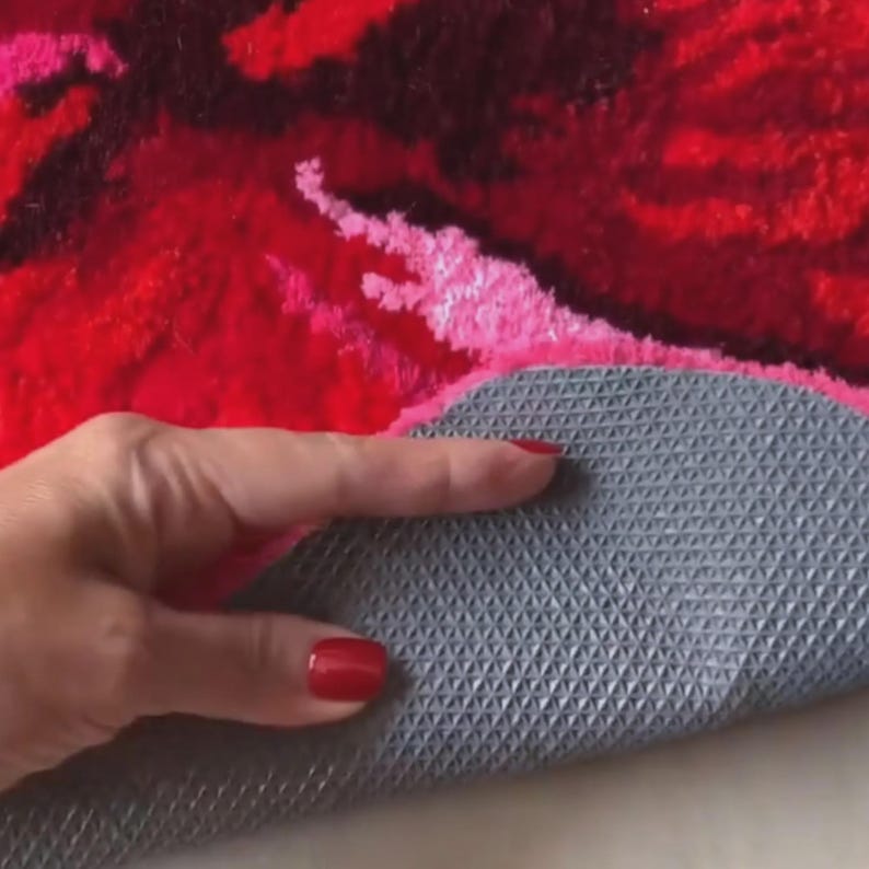 May include: A close-up of a red, pink, and black floral rug. The rug's underside is gray with a textured, non-slip pattern. A hand with red nail polish is holding the rug.