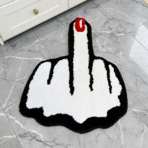 May include: A novelty rug in the shape of a hand giving the middle finger. The rug features a white central area, a black border, and a red fingernail. It is placed on a marble-effect tiled floor.