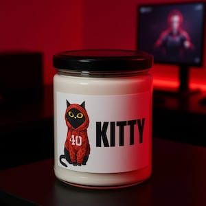 Könnte beinhalten: Eine weiße Kerze in einem Glas mit schwarzem Deckel. Das Etikett zeigt eine Cartoon-Katze in einem roten Kapuzenpullover mit der Nummer 40 und dem Wort "KITTY" in großen schwarzen Buchstaben. Der Hintergrund ist verschwommen rot.