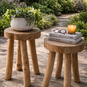 May include: Two small, natural wood stools with three legs each. One stool holds a white speckled planter with greenery. The other holds books, eyeglasses, and a lit candle. The scene is set outdoors on a stone patio.