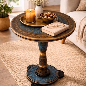May include: Round, ornate side table with a blue and gold patterned top and base. A lit candle, small vase of flowers, bowl of nuts, and a book with glasses rest on the table. The table is 24 inches in diameter.