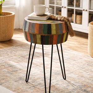 May include: A round side table with a colorful, striped design on the tabletop and sides. The table has black hairpin legs and is topped with a white mug and a stack of books. A decorative wooden bead garland rests on the table.