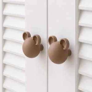 May include: Two brown, bear-shaped cabinet knobs are mounted on a white, louvered door. The knobs have a rounded shape with two small, curved ears. The door is part of a larger cabinet or piece of furniture.