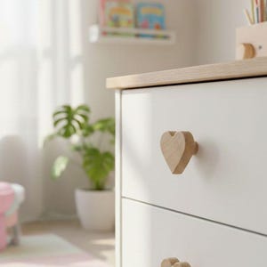 Puede incluir: Cómoda blanca de dos cajones con tiradores de madera en forma de corazón. La parte superior de la cómoda es de madera clara. La cómoda está en una habitación luminosa con una planta y una ventana.