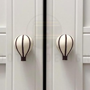 May include: Two off-white cabinet knobs shaped like hot air balloons, each featuring vertical dark brown stripes and a dark brown base. The knobs are mounted on a light gray cabinet door with vertical paneling and a central vertical gap.