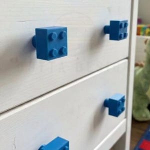 May include: A white wooden chest of drawers featuring four blue drawer knobs shaped like rectangular building blocks. Each knob has four circular studs on the front face. The chest has two drawers, with two blue block-style knobs attached to each drawer front.