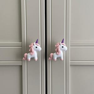 May include: Two unicorn-shaped cabinet knobs. The knobs are white with pink manes, tails, and hooves. They have purple horns and are mounted on a light gray cabinet door. The knobs are designed to add a whimsical touch to furniture.