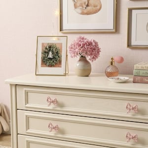 May include: A cream-colored wooden dresser featuring four drawers with light pink bow-shaped knobs. The dresser is topped with a small vase of pink hydrangea flowers, a glass perfume bottle, a small white dish, and three stacked books.