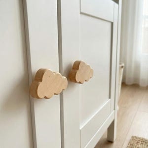 May include: White cabinet with two cloud-shaped wooden knobs. The knobs are made of light-colored wood and are attached to the cabinet doors. The cabinet has a simple, modern design, and the knobs add a whimsical touch.